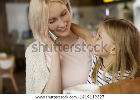 Side view of cheerful girl hugging and looking at mother while she talking by phone in cafe. Attractive woman calling husband while sitting with daughter in restaurant. Concept of parental love.