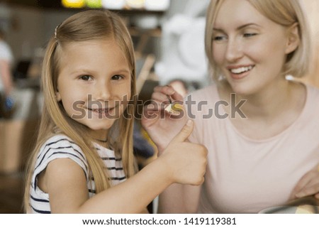 Front view of little girl looking at camera and showing sign ok while mother feeding her with delicious cheesecake in cafe. Happy family having lunch and enjoying dessert. Concept of good taste.