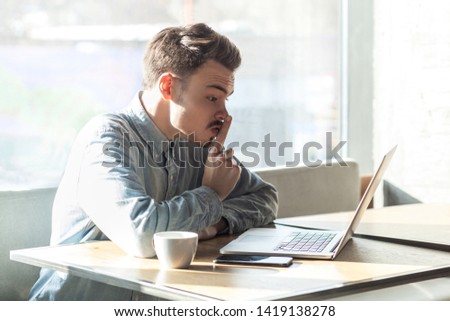 Be quiet please! Side view portrait of severe bearded young freelancer in blue shirt are sitting in cafe and making video call on laptop, lean finger on mouth showing silence sign to companion.