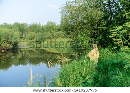 Fisherman male catch fish on river coast in grass, man in green clothes and hat holds fishing-rod, view from back, horizontal stock photo image