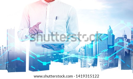 Unrecognizable businessman in white shirt using tablet computer over cityscape background with double exposure of graphs. Concept of trading and hi tech. Toned image