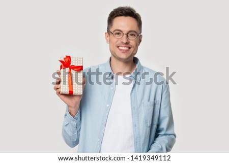 On grey studio wall smiling man in glasses looking at camera holds gift box with red ribbon presenting new product, advertise discount card eyewear store, advice how make happy girlfriend for holiday