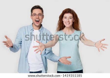 Excited couple happy smiling red-headed girl guy in glasses looks at camera raise hands make embrace cuddle gesture welcome friend or greeting client nice to meet you concept studio image on grey wall Royalty-Free Stock Photo #1419351185