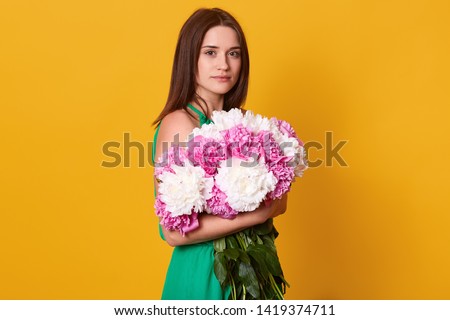 Studio shot of beautiful brunette female embracing big bouquet with pink and white peonies, stylish woman with flowers, has calm facial expression, posing isolated over yellow studio background.