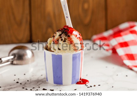  vanilla ice cream in ceramic bowl with Strawberry syrup and Chocolate noodles on marble table with scoop and spoon for ice cream on wooden backdrop