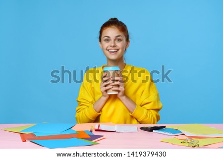 A woman with a cup of coffee in her hand a yellow sweater a pink table and multi-colored folders                