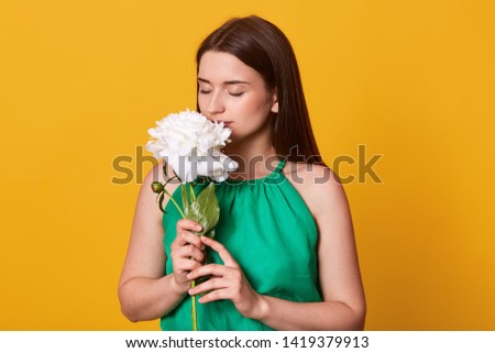 Indoor shot of romantic sweet female standing isolated over bright yellow background in studio, wearing green summer dress, closing her eyes, holding white peony, smelling it, being satisfied.