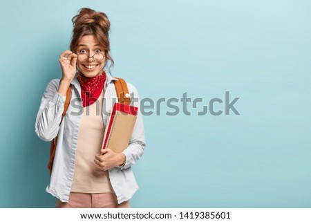 Indoor shot of happy good looking young college student wears optical round glasses, carries textbook and notepad, ready for classes, stands against blue wall with empty space. Studying concept