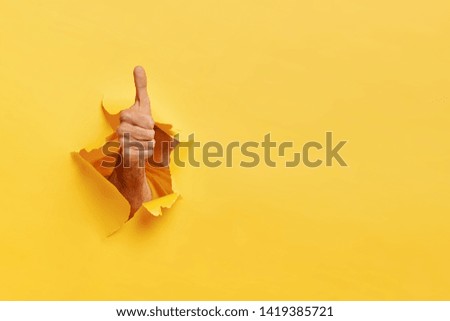 Unrecognizable man shows like gesture through torn yellow wall, keeps thumb up, says you are best, demonstrates approval sign, recommends something. Copy space aside for your advertising content Royalty-Free Stock Photo #1419385721