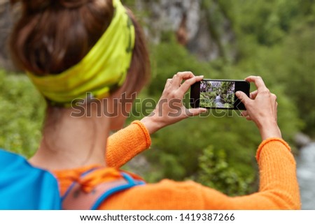 Backwards shot of woman tourist makes photo of river in ravine on smartphone device for posting in social networks, makes video for chanel, carries backpack, amazing nature view. Place for inscription
