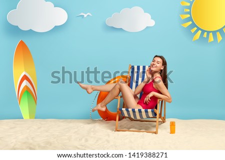 Slim good looking female rests in deckchair at beach, shows slender legs, wears red bikini, has glad look, charming smile, bottle of sunscreen on sand, surfboard has unforgettable summer holiday