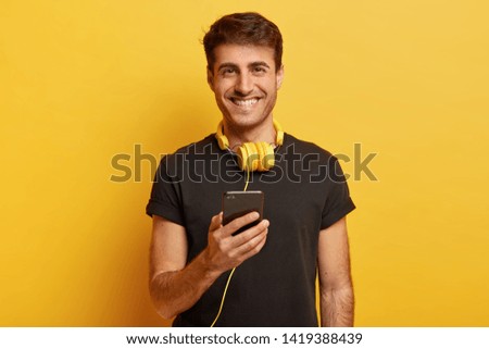 Portrait of happy Caucasian man enjoys fantastic sound and quality of headphones, holds modern cell phone, listens music, smiles joyfully, isolated over yellow background. People and pastime concept
