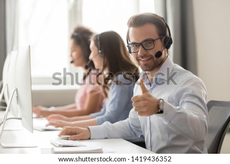 Smiling male employee wearing headset sit at shared office desk working at pc show thumb up, glad millennial man worker call center agent recommend service, consult clients on computer online