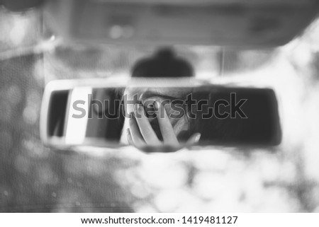 Black and white art picture of young woman covering her face hands in the car rear view mirror. 