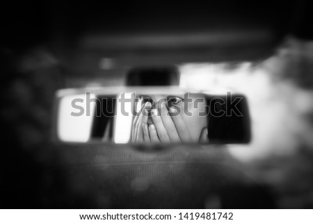 Black and white art picture of young woman with frightened eyes covering her face with hands in the car rear view mirror. 