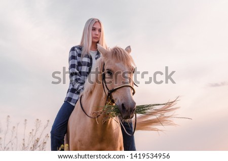 young blonde girl dressed in blue jeans and a plaid shirt sits on a horse at the ranch closeup