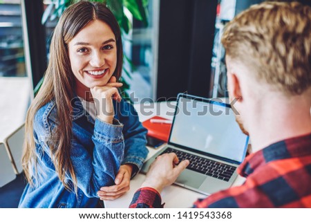 Cheerful woman dressed in casual apparel looking at camera while male friend near typing text information to laptop application for making advertising online, concept of technology and communication