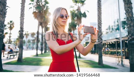 Trendy blond woman in sunglasses and red overall using mobile phone and taking photo of Barcelona standing in back lit