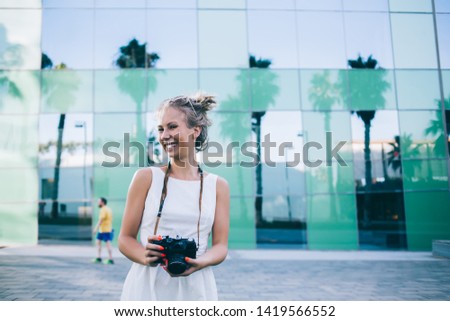 Beautiful young blond woman in white dress and sunglasses holding modern camera and laughing at camera standing on street of Barcelona city