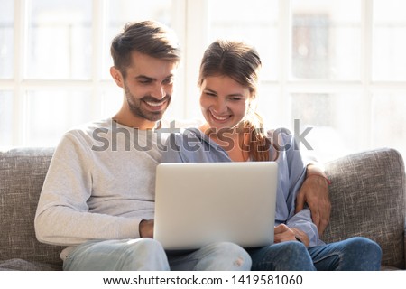 Happy young couple sit relaxing on couch hugging watching funny video on laptop, smiling loving man and woman embrace cuddle on sofa at home using computer, laugh surfing internet together