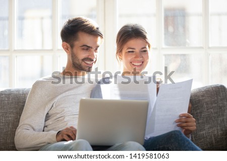 Smiling husband and wife sit on couch using laptop taking care of utility bills and house maintenance documents, happy young couple relax on sofa read paperwork, pay bank credit online via internet Royalty-Free Stock Photo #1419581063