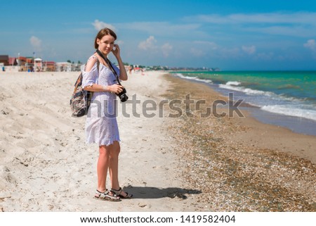 Woman photographer at the sea. A middle-aged woman in a white dress with a camera goes along the coast. Summer tourism, photographing attractions on vacation.
