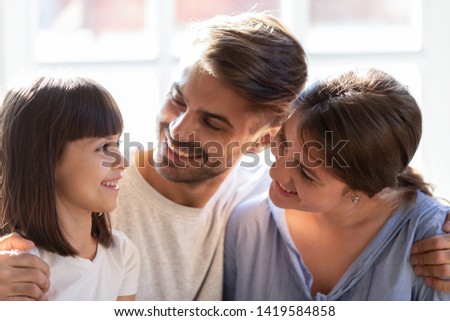 Happy young family with little preschooler kid cuddle relaxing at home together, smiling mom and dad look at cute small daughter enjoy tender sweet time, excited parents hug spend weekend with child