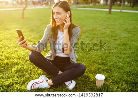 Image of beautiful stylish woman sitting on green grass with a phone in hand and coffee. She is talking on the phone through wireless headphones. Sunset light. Lifestyle concept.
