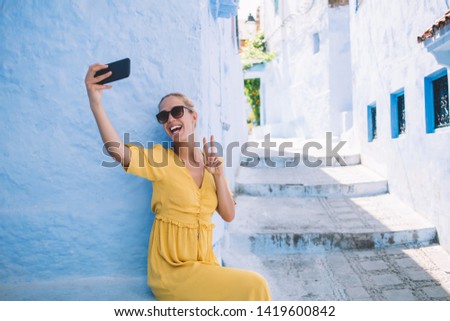 Happy woman in trendy sunglasses enjoying time for shooting peace video and create social web publication for web blog, cheerful female tourist clicking selfie pictures via cellphone front camera