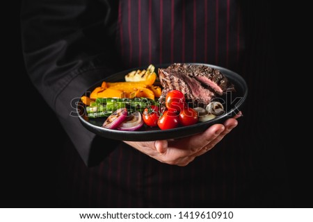 Grilled and sliced beef steak with grilled vegetables served on black plate on black background presentation in chef hands.