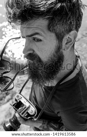 Man with beard and mustache puts on sunglasses, water surface on background. Hipster on strict face holds old fashioned camera. Tourist photographer concept. Guy shooting nature near river or pond.