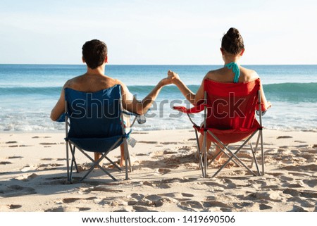 Rear View Of Young Couple Sitting On Chairs Holding Each Other's Hands Looking At Sea