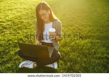 Image of beautiful stylish woman sitting on green grass with laptop and coffee in the hand. She is talking on the phone through wireless headphones. Sunset light. Lifestyle concept