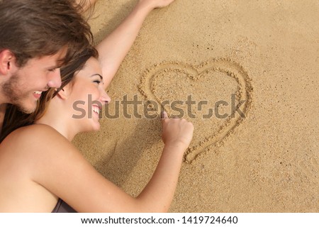 Happy couple in love lying and drawing a heart on the sand of the beach