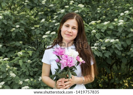 Beautiful girl in a white T-shirt with peonies in their hands against the background of green bushes