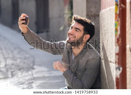 Excited trendy young man taking selfie or blogging on smart mobile phone on holidays in european city street or university campus. Holidays and technology Connection and social media concept.
