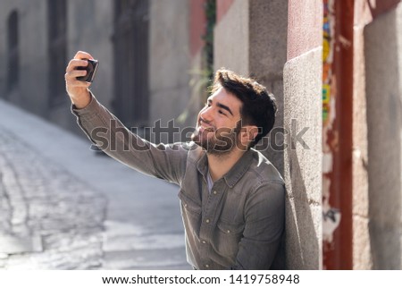 Excited trendy young man taking selfie or blogging on smart mobile phone on holidays in european city street or university campus. Holidays and technology Connection and social media concept.