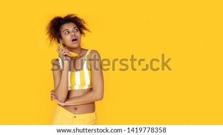Curious young african woman holding banana look aside at copy space, black teen girl pretending talk on fruit phone or listen gossip isolated on summer studio background, banner website design