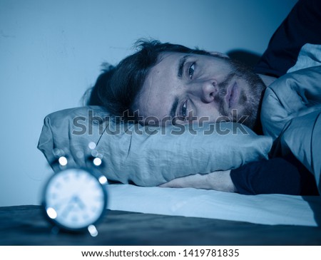 Sleepless and desperate young caucasian man awake at night not able to sleep, feeling frustrated and worried looking at clock suffering from insomnia in stress and sleeping disorder concept. Royalty-Free Stock Photo #1419781835
