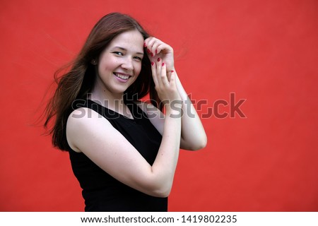 Photo portrait of a beautiful young pretty girl with dark red hair on a red background in a black jacket. Standing right in front of the camera in different poses, smiling, talking. Made in a studio.