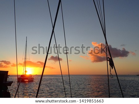 Through sailboat rigging at a catamaran at anchor with yellow-orange sunset glowing behind, pink billowing cloud light blue sky over calm ocean water. Beautiful background with room for copy space.