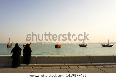 Two women in traditional Arab dress taking photos of Doha bay with traditional boats. High Resolution Photography.       
