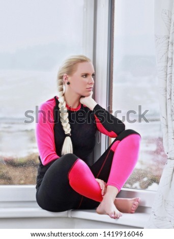 Sporty European woman with long blond hair dressed warm pink clothing sitting on the window on the ski resort on background looking away. Leisure activities and winter sports concept.