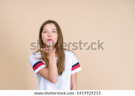 Front view of happy adult girl looking at camera and sending air kiss from open palm standing isolated on pastel beige background with copy space