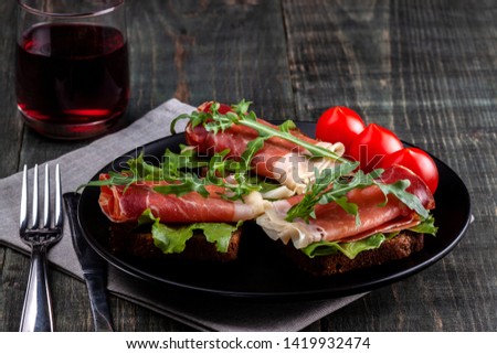 On the table there is a dish with jamon sandwiches, tomato, greens and a glass of red wine