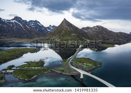 A artistic bridge connecting an island in the mountains over blue ocean water with clouds reflections and snow covered mountain summits in the background in Lofoten Islands, Norway