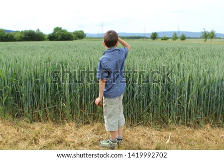 boy in a denim shirt and shorts stands at the edge of a green field with a rich harvest of wheat in the ears ripening in the fields, shows his right hand to distant mountains