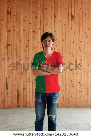 Man wearing Portugal flag color shirt and cross one's arm on wooden wall background, 2:3 vertically striped of green and red with coat of arms of Portugal. 