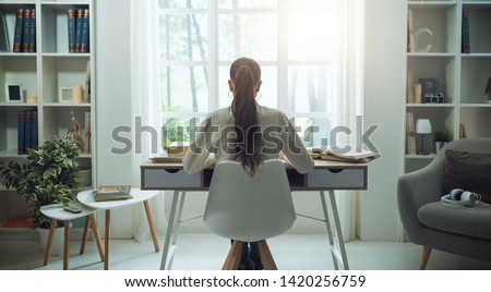 Young woman sitting at desk and studying at home, she is reading books, back view Royalty-Free Stock Photo #1420256759