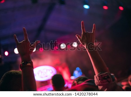Young teenager girl raised up two hands with sign of the horns gesture supporting favorite rock band on the night concert  